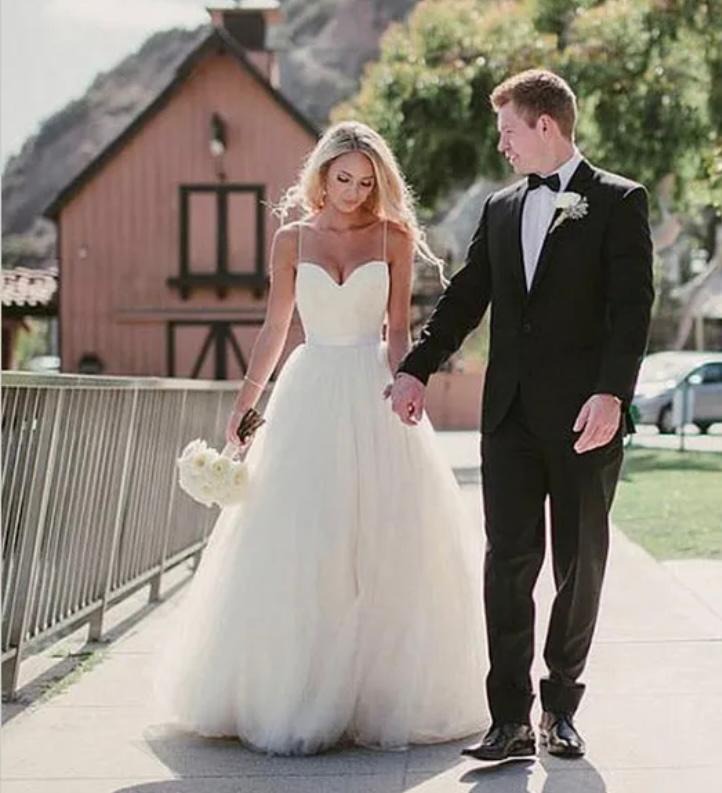 Para młoda, blondynka w sukni z tiulu i mężczyzna w czarnym smokingu, spacerują trzymając się za ręce na tle rustykalnego budynku z czerwonej cegły.