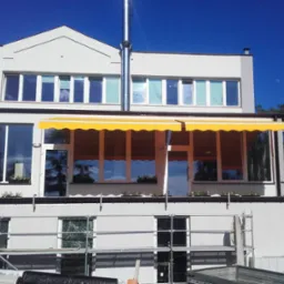 Żółta markiza nad balkonem budynku, widoczne rusztowania, okna z odbiciami drzew, niebieskie niebo w tle.