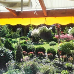 Żółta markiza z falbaną zamontowana nad tarasem, z wiszącymi doniczkami z białymi kwiatami i widokiem na zadbany ogród z różnorodną roślinnością.