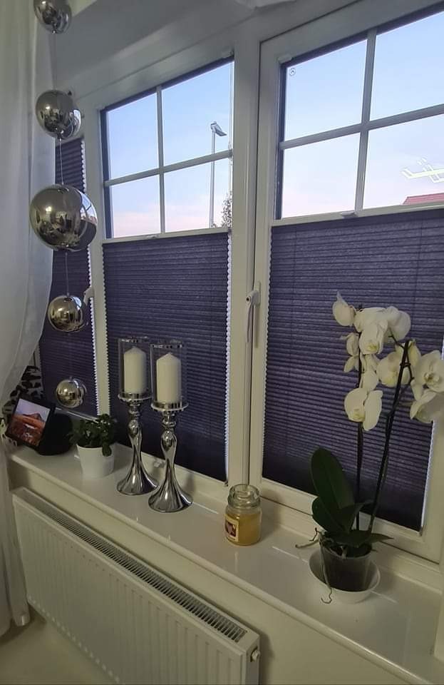 Dwa okna z częściowo zasłoniętymi, ciemnymi panelami, dekoracje okienne: srebrne kule, świeczniki, kwiaty, słoik na parapecie.