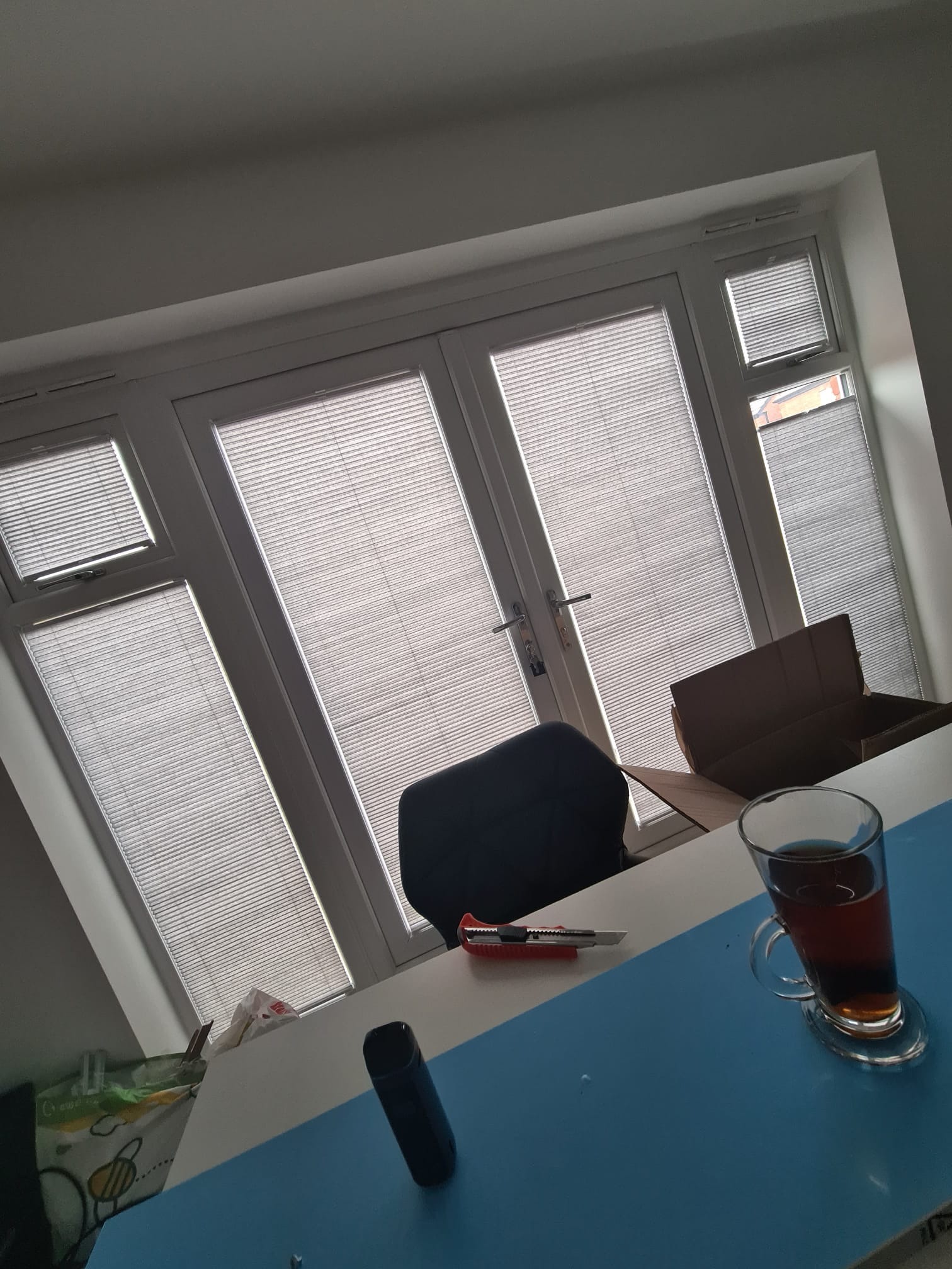 Okna dachowe z zamontowanymi plisami w kolorze szarym, widok od dołu, na pierwszym planie biurko z kubkiem herbaty i nożem introligatorskim.