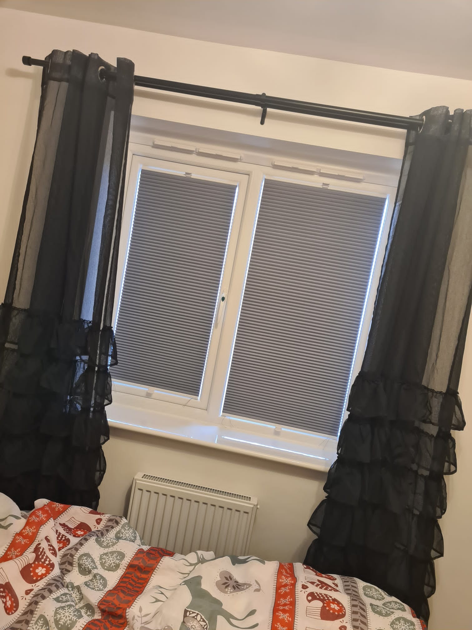 Pokój z oknem zasłoniętym szarymi plisami, po bokach czarne, prześwitujące zasłony z falbanami, widoczny fragment łóżka z pościelą w świąteczne wzory.