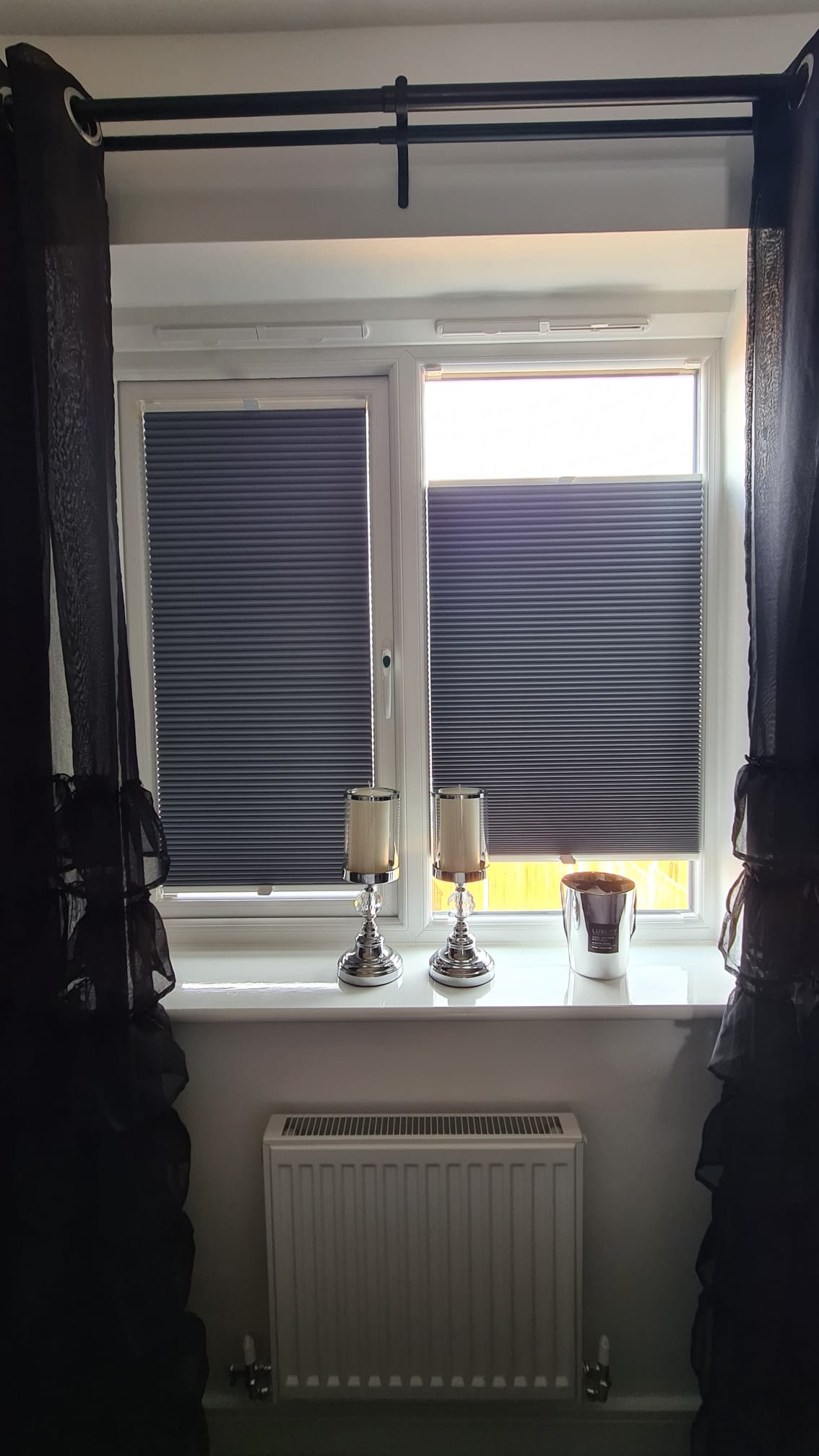 Okna z szarymi plisami, ozdobione świecznikami i czarnymi zasłonami, widok na grzejnik pod parapetem.