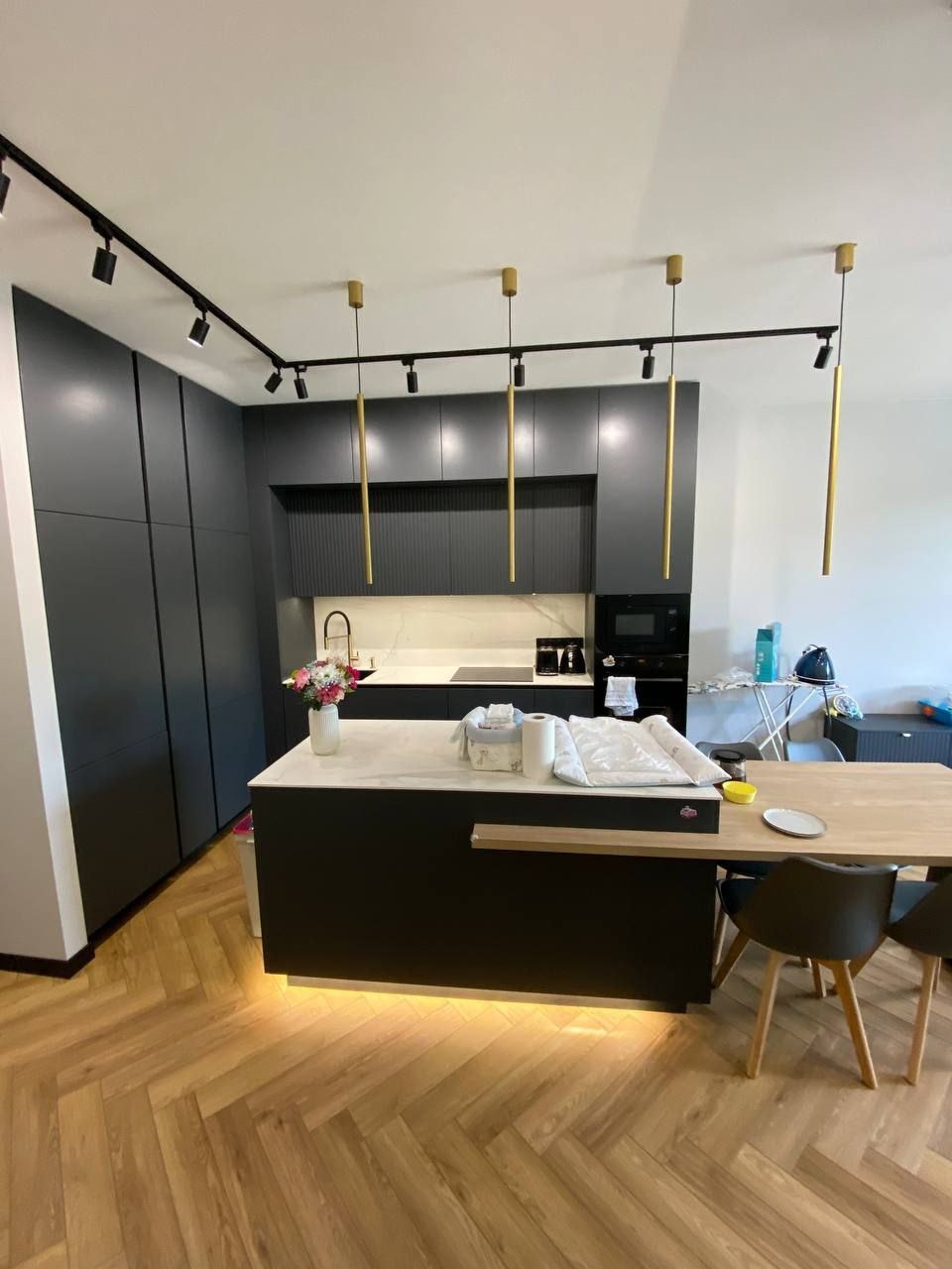 Nowoczesna kuchnia z ciemnymi szafkami, marmurowym blatem, wyspą z podświetleniem LED i parkietem ułożonym w jodełkę.