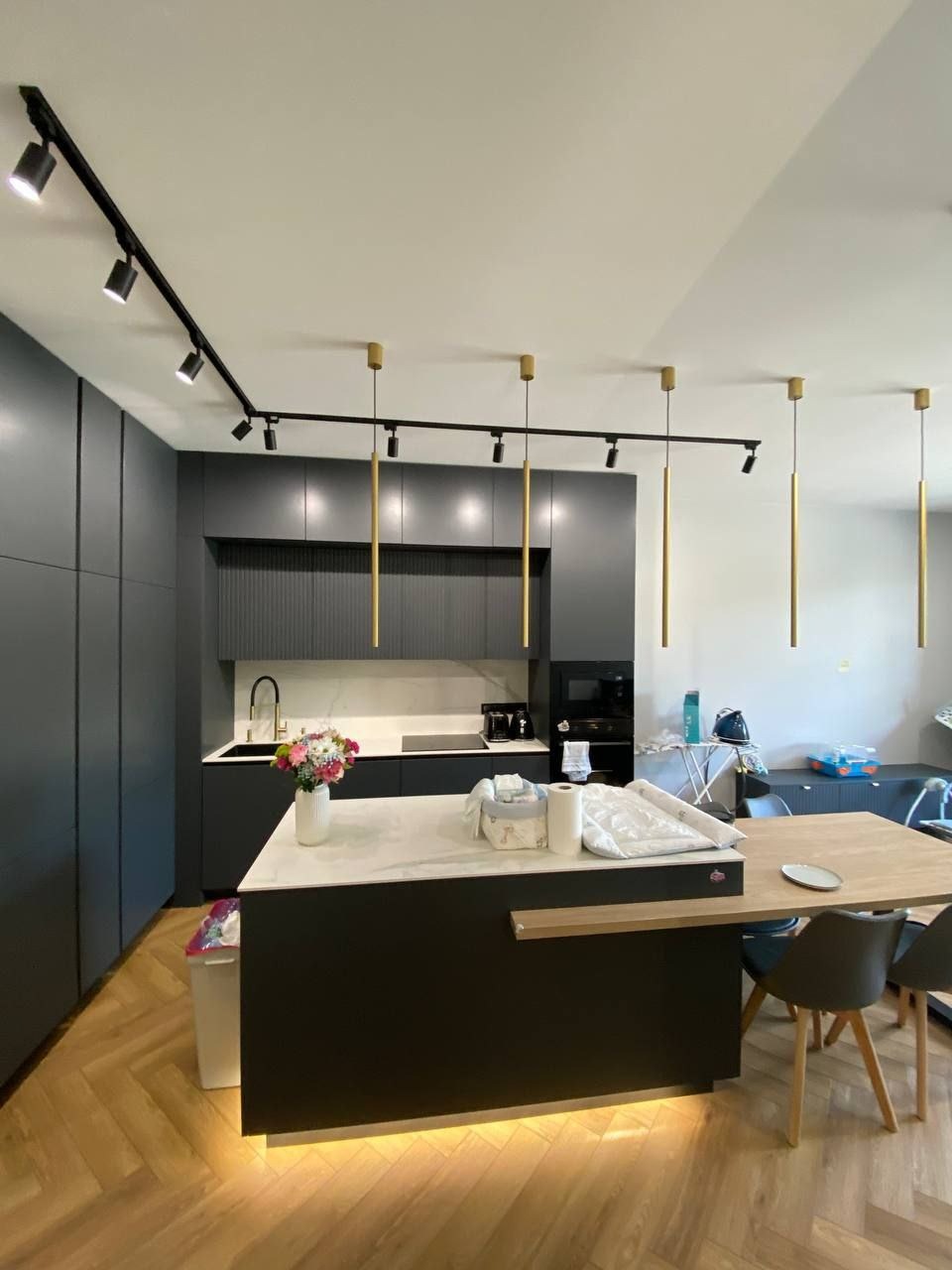 Nowoczesna kuchnia w odcieniach szarości z drewnianą podłogą w jodełkę, wyspą z białym blatem i złotymi lampami wiszącymi.