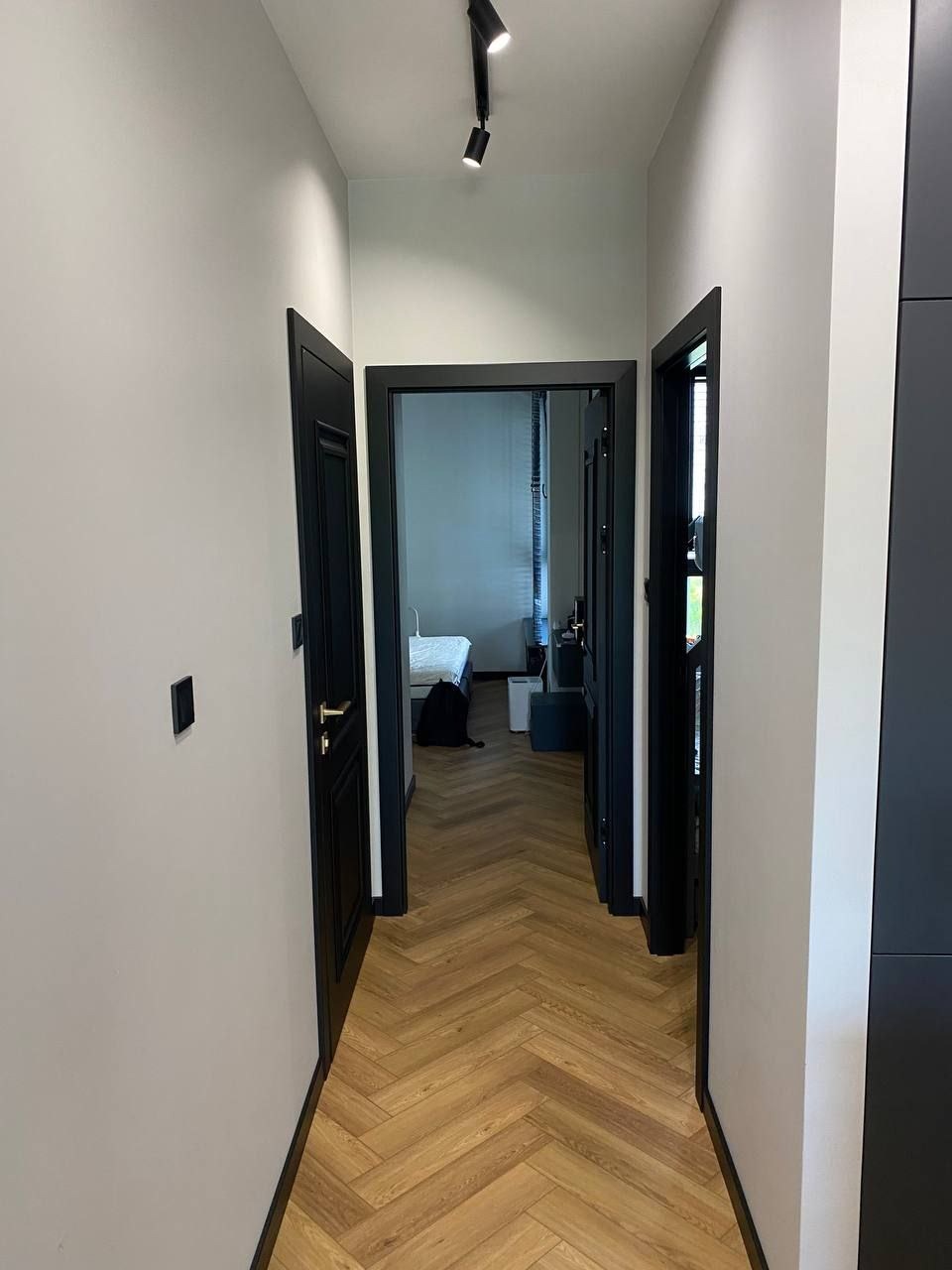 Korytarz z parkietem ułożonym we wzór jodełki, czarne drzwi z złotymi klamkami, białe ściany i oświetlenie punktowe na suficie.