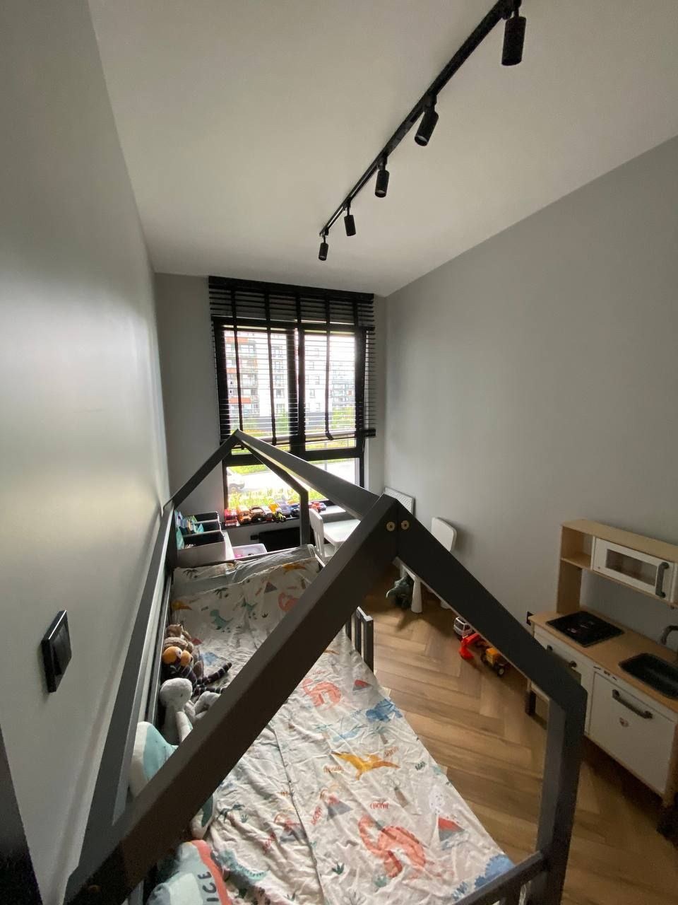 Pokój dziecięcy z łóżkiem w kształcie domku, drewnianą podłogą w jodełkę i szarymi ścianami, oświetlony reflektorami sufitowymi.