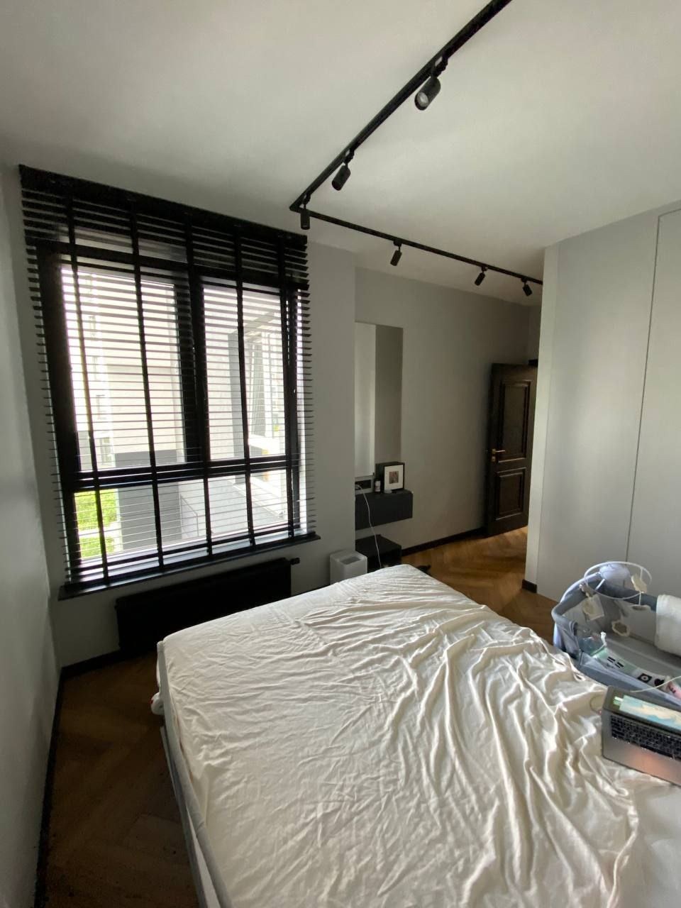 Jasne wnętrze sypialni z drewnianą podłogą w jodełkę, łóżkiem z białą pościelą, czarnymi żaluzjami w oknie i reflektorami na suficie.