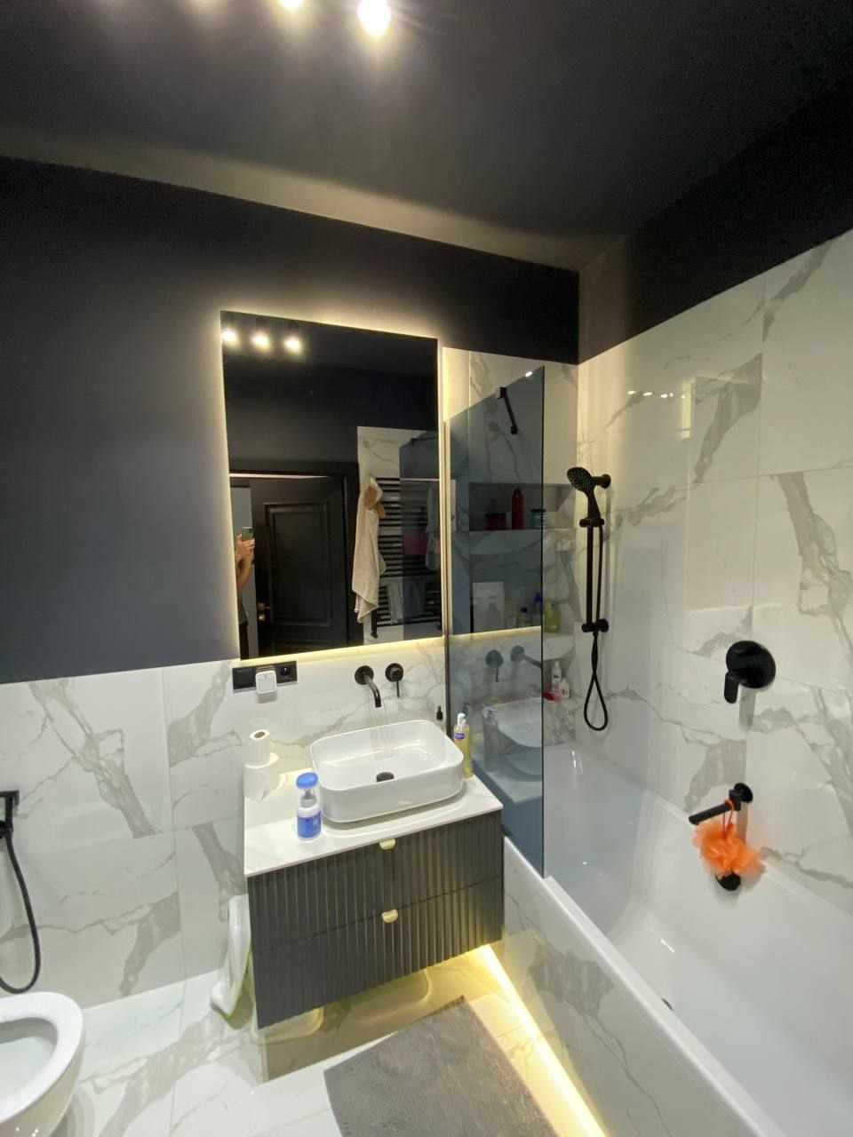 Nowoczesna łazienka z czarnym sufitem, białymi marmurkowymi płytkami, podświetlanym lustrem i szarą szafką pod umywalką oraz czarną armaturą.