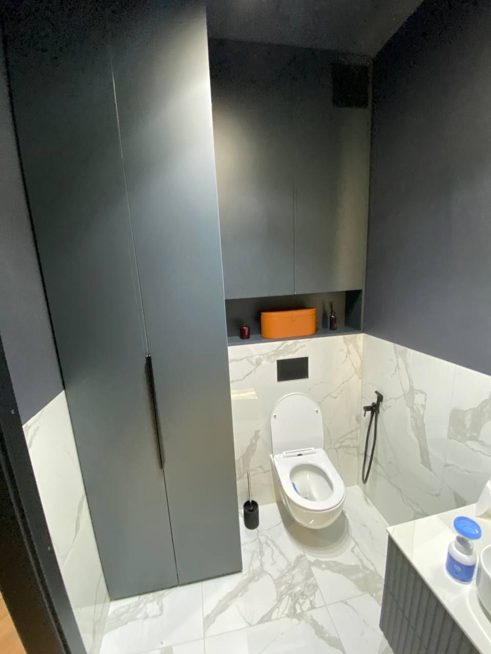 Nowoczesna toaleta w odcieniach szarości i bieli z marmurową podłogą, zabudową meblową pod sufit, wiszącą muszlą klozetową i minimalistyczną armaturą natryskową.