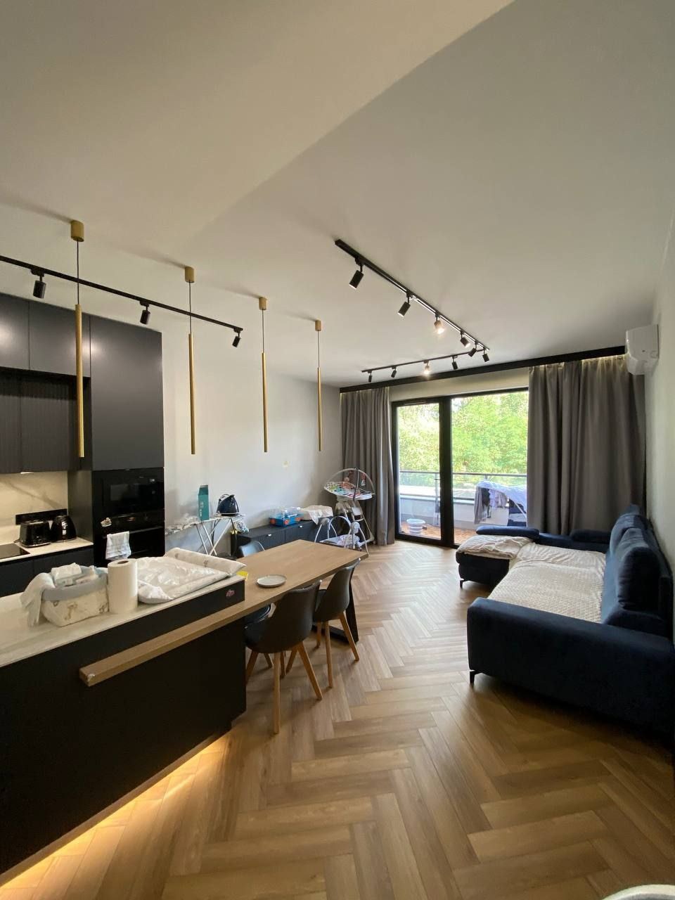 Salon z aneksem kuchennym, parkiet ułożony w jodełkę, czarne oświetlenie szynowe i wiszące lampy w kolorze złotym, granatowa sofa i balkon z widokiem na zieleń.