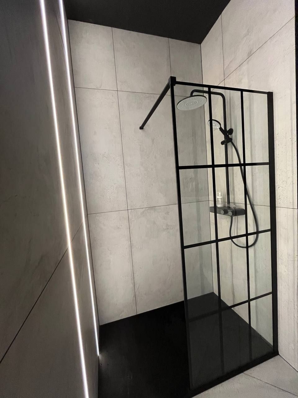 Nowoczesna kabina prysznicowa z czarnymi ramami i deszczownicą, zamontowana w łazience w Warszawie z szarymi płytkami i czarną podłogą, oświetlona pionowym paskiem LED.