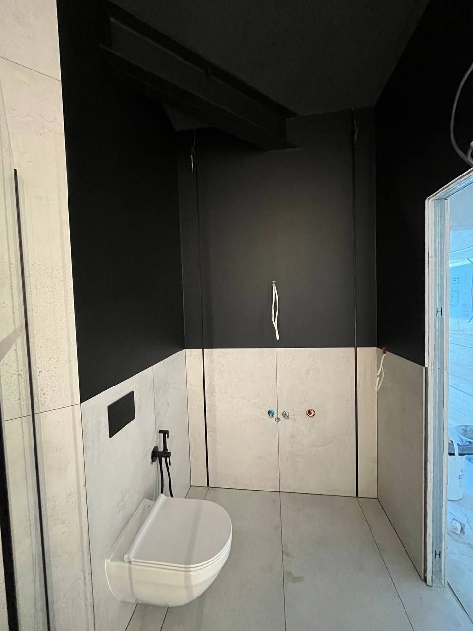 Nowoczesna łazienka w trakcie wykańczania: białe płytki imitujące kamień, czarne ściany do połowy wysokości, podwieszana toaleta z bidetką, widoczne przyłącza wodne i fragment otwartego prysznica.