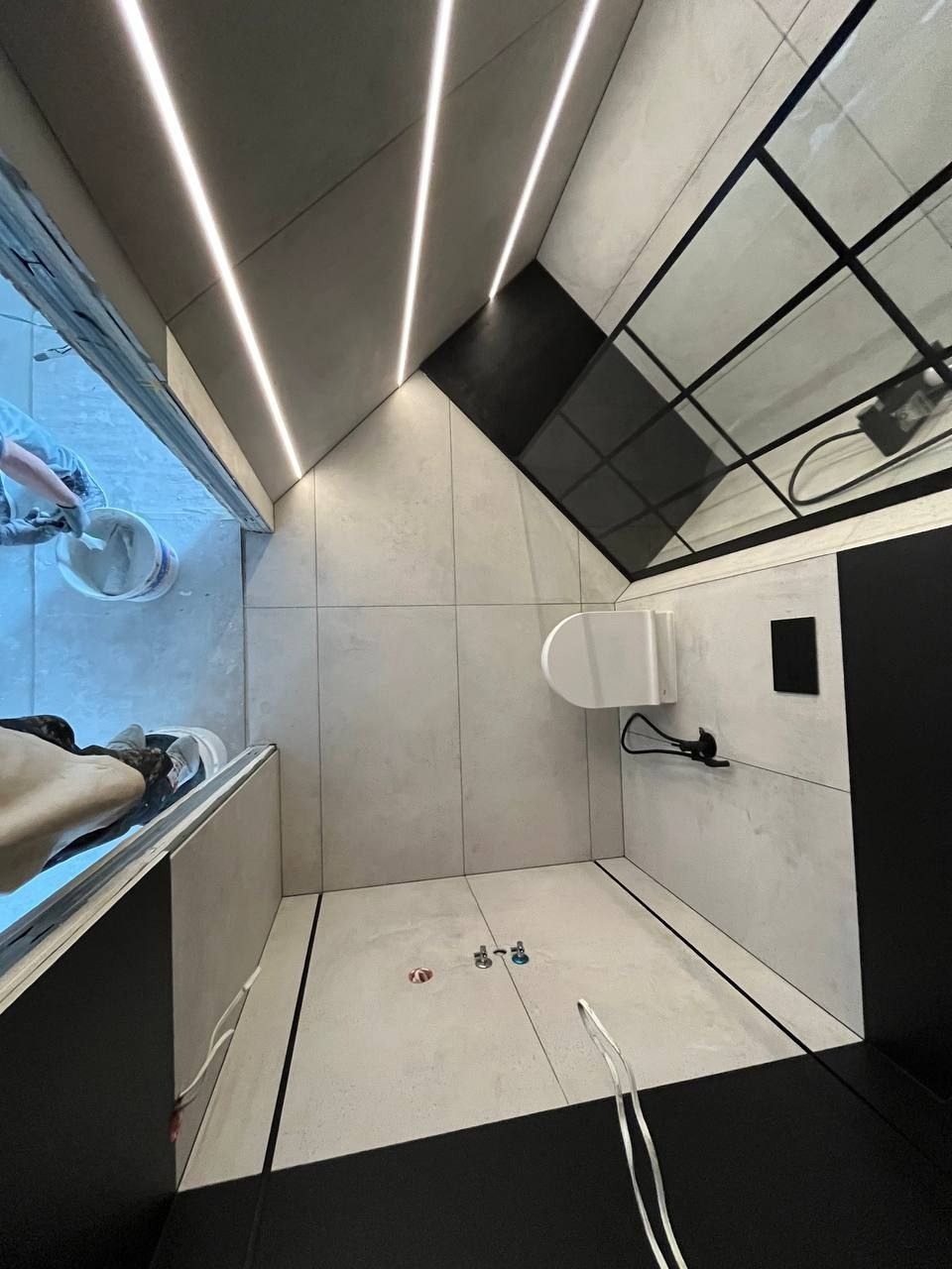 Widok z góry na nowoczesną łazienkę w trakcie wykańczania: szare płytki, czarne akcenty, oświetlenie LED w suficie, widoczna osoba przygotowująca zaprawę w wiadrze, niedokończona kabina prysznicowa.