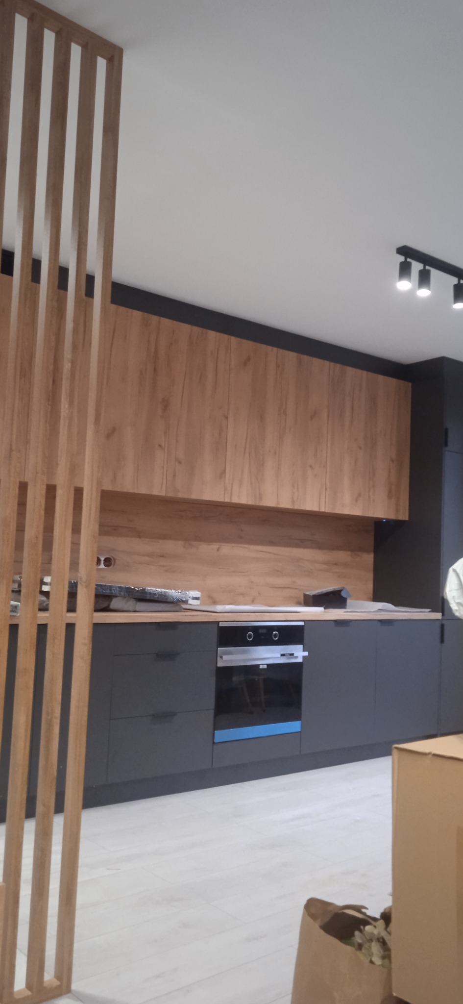 Nowoczesna kuchnia z drewnianymi szafkami górnymi i szarymi dolnymi, widoczna drewniana ażurowa ścianka działowa, oświetlenie punktowe na suficie.