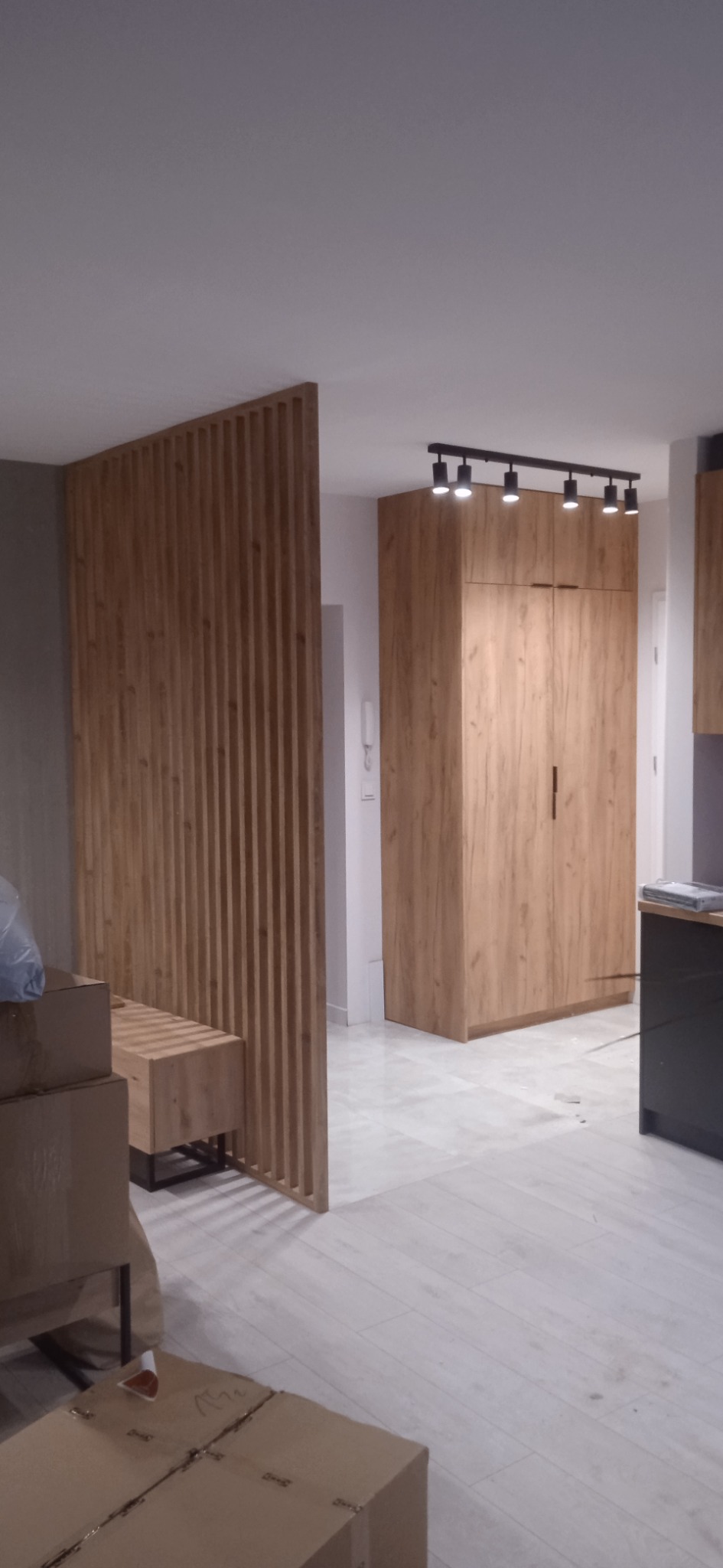 Drewniana ścianka działowa z pionowych listew, szafa w zabudowie z oświetleniem punktowym, podłoga z jasnych paneli, kartony w tle. Wnętrze w trakcie wykańczania.