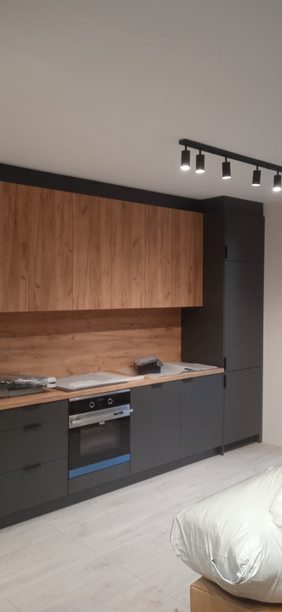 Nowoczesna kuchnia z szarymi szafkami dolnymi i drewnopodobnymi górnymi, oświetlenie punktowe na suficie, blaty zabezpieczone folią ochronną, częściowo widoczny sprzęt AGD.