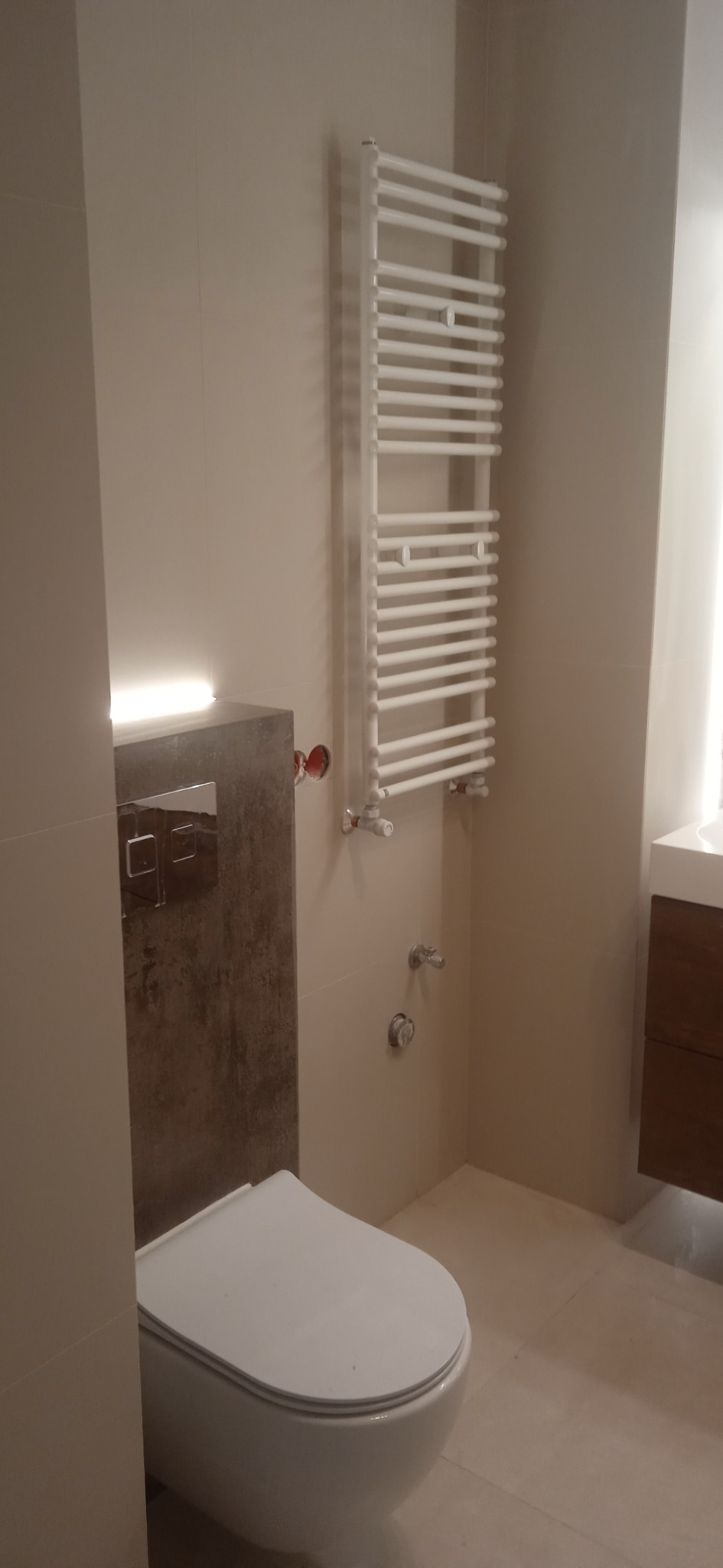 Nowoczesna łazienka z podwieszaną toaletą, grzejnikiem drabinkowym i umywalką z szafką w odcieniach beżu i brązu.