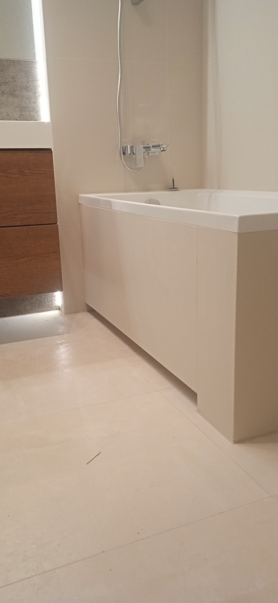 Minimalistyczna, wykończona łazienka w Warszawie z wanną obudowaną panelami w kolorze złamanej bieli, chromowaną armaturą i drewnianą szafką pod umywalką z podświetleniem LED.