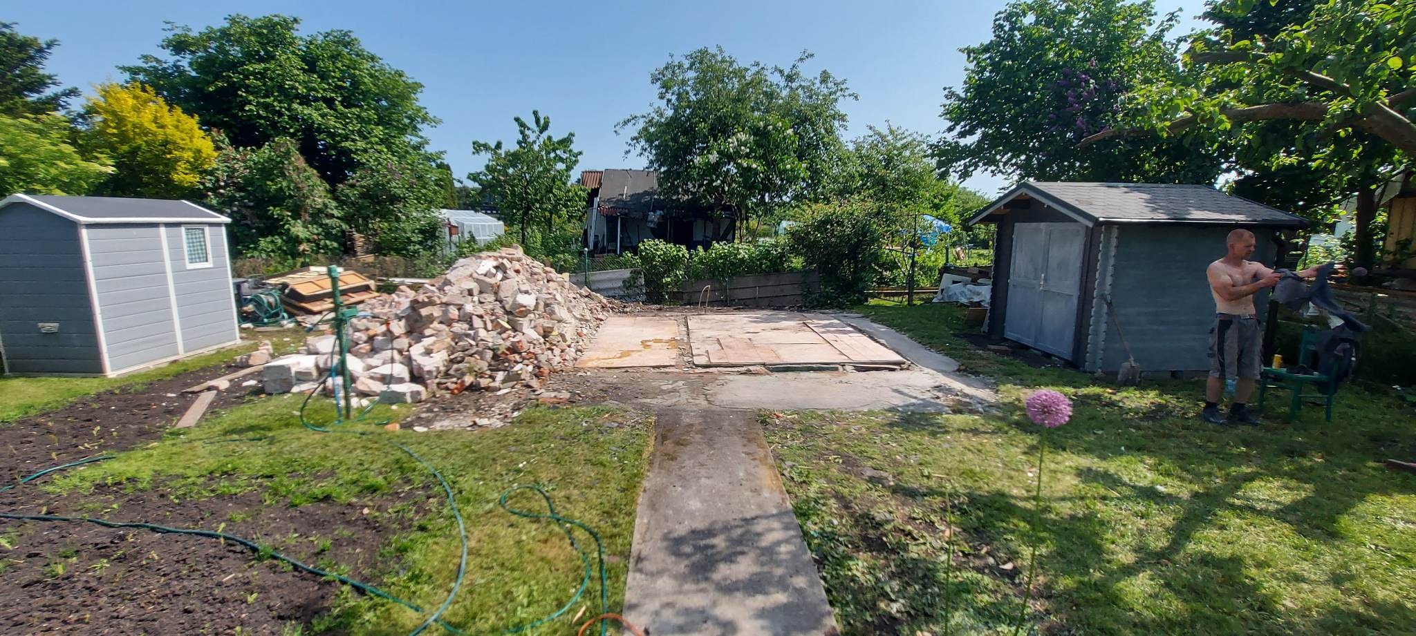 Teren po rozbiórce budynku z stertą gruzu, przygotowany pod budowę, z widocznym zarysem fundamentów i dwoma blaszakami w tle, jeden szary i jeden drewniany, oraz mężczyzną bez koszulki.