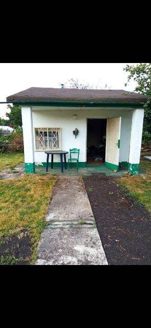 Niewielki, parterowy budynek z płaskim dachem, pomalowany na biało z zielonymi akcentami, widoczny z zewnątrz, z otwartymi drzwiami, przez które widać wnętrze, obok wejścia stoi stolik i krzesło.