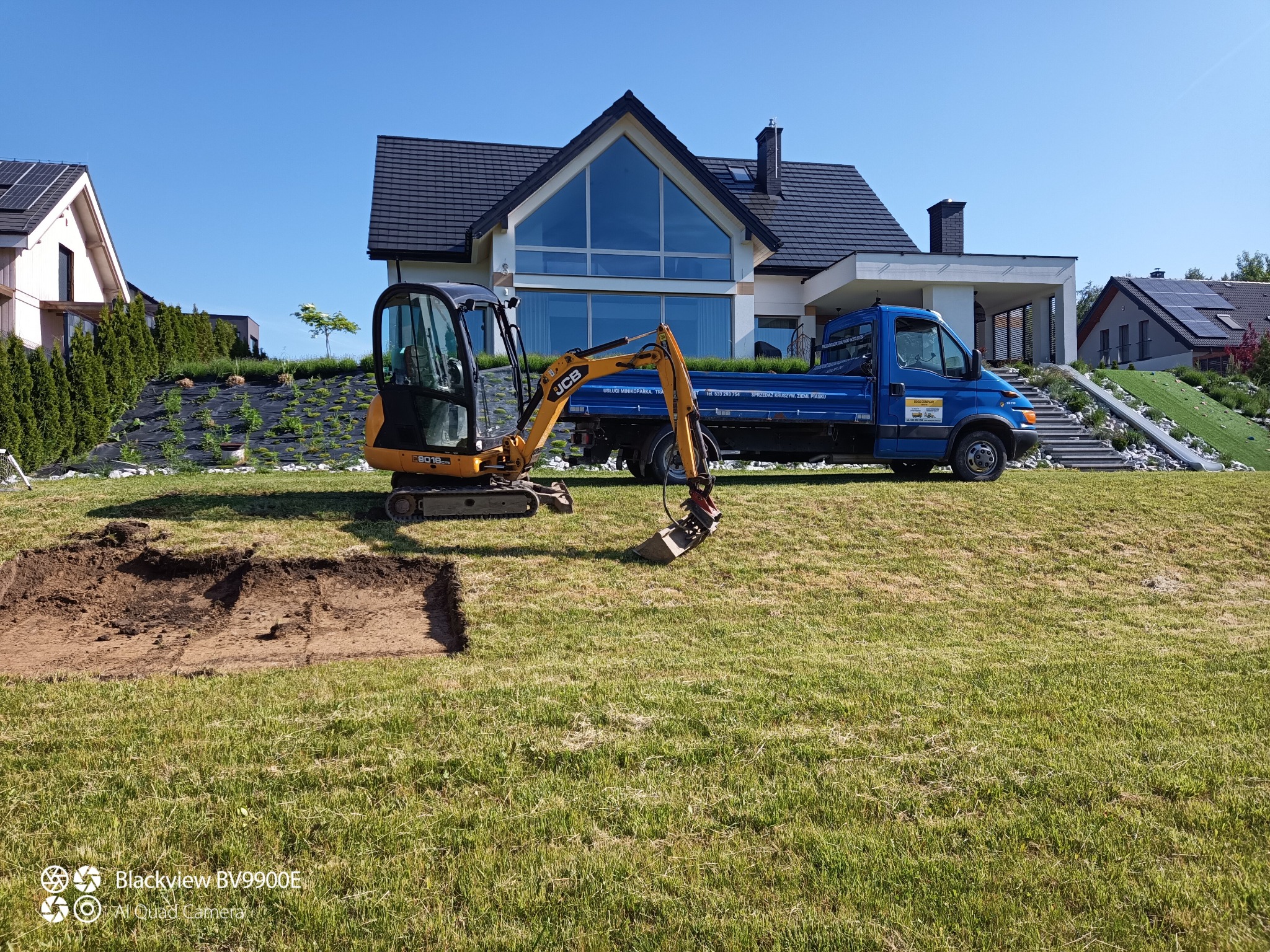 Mini koparka JCB wykonuje wykop w trawniku na tle nowoczesnego domu z dużym oknem i niebieskiej ciężarówki.
