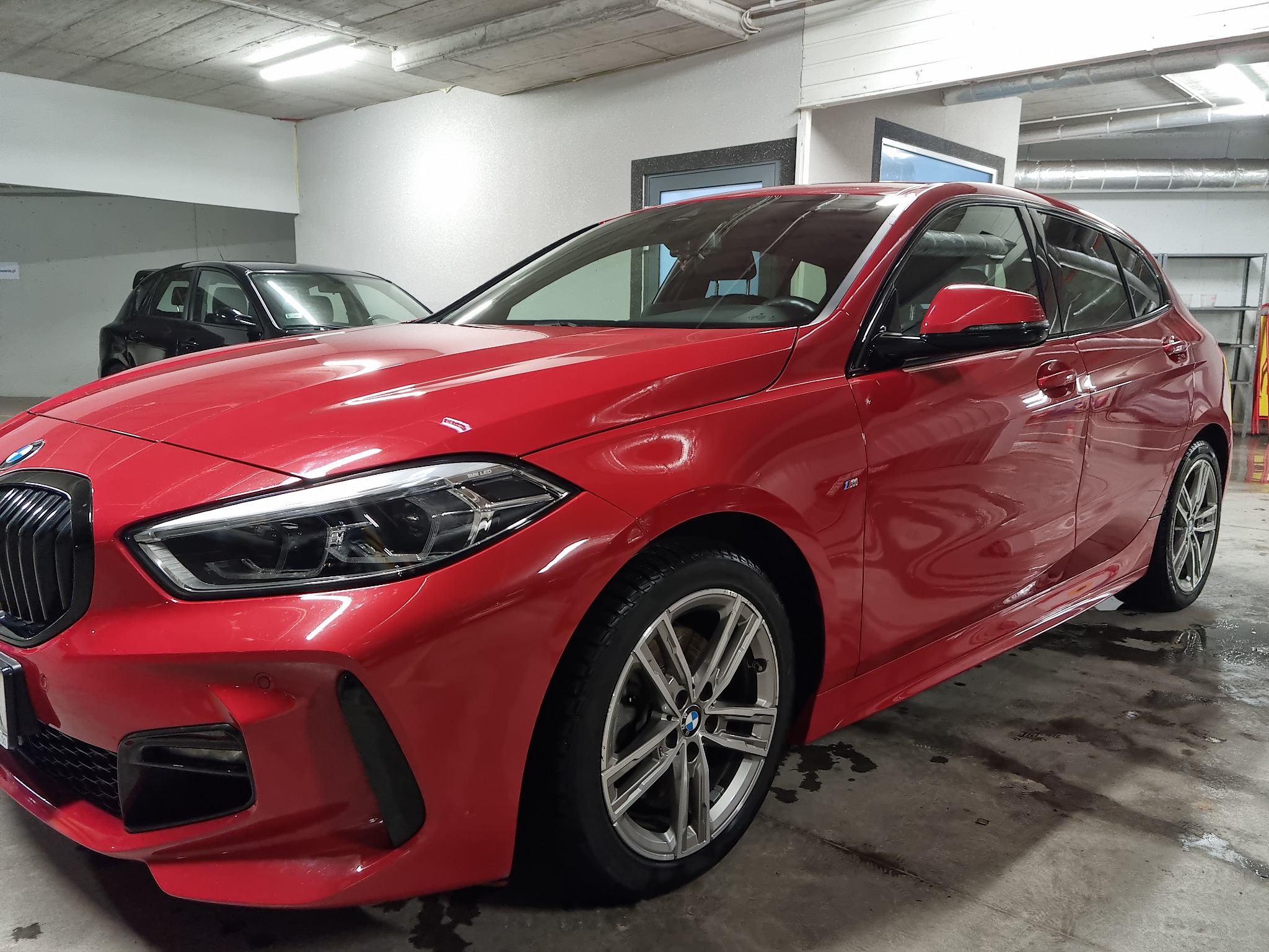 Czerwony hatchback BMW po umyciu, widoczne krople wody na karoserii, zaparkowany w garażu podziemnym.