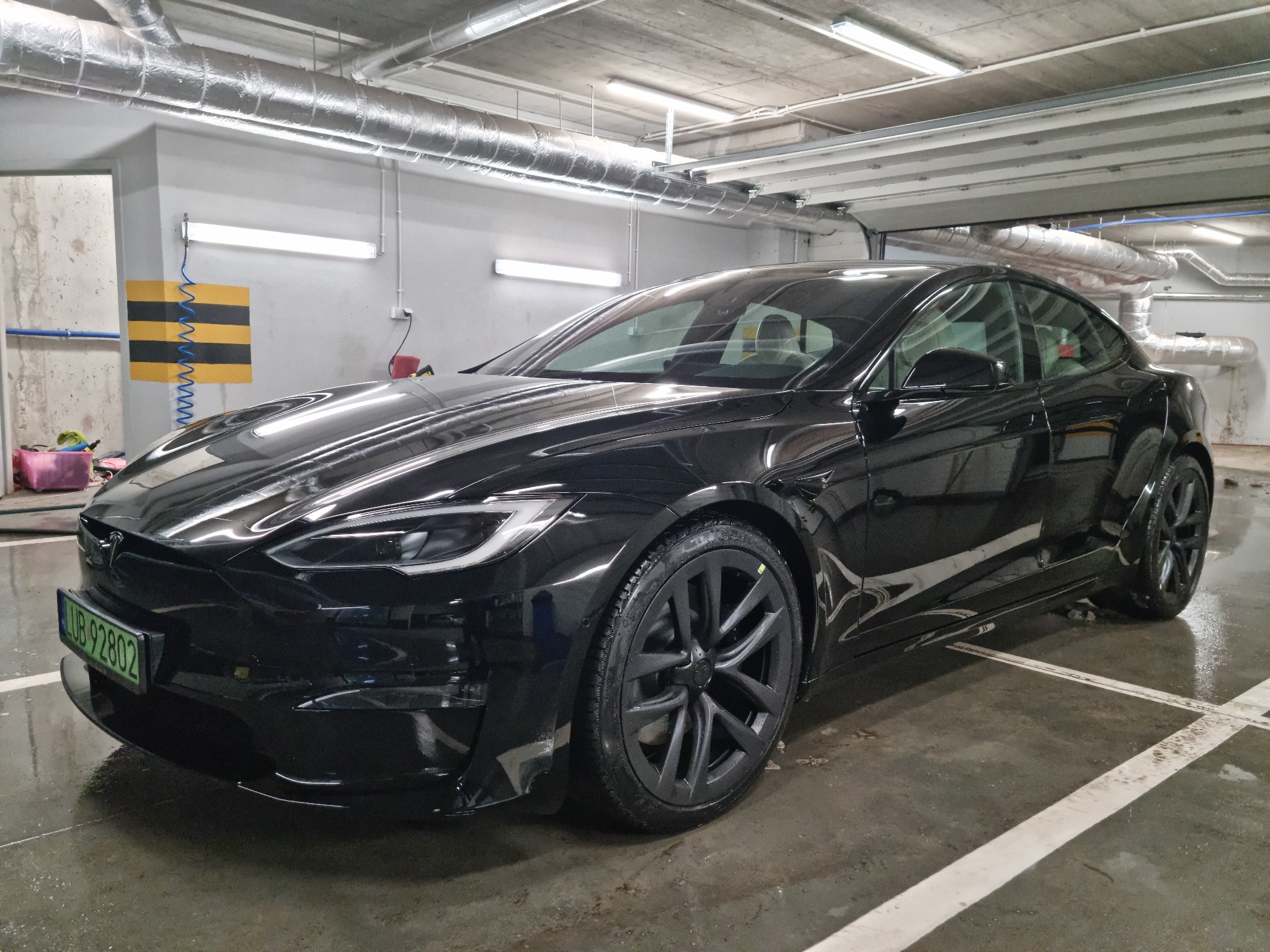 Błyszczący, czarny samochód elektryczny Tesla Model S po umyciu, zaparkowany w garażu podziemnym z widocznymi instalacjami wentylacyjnymi pod sufitem.