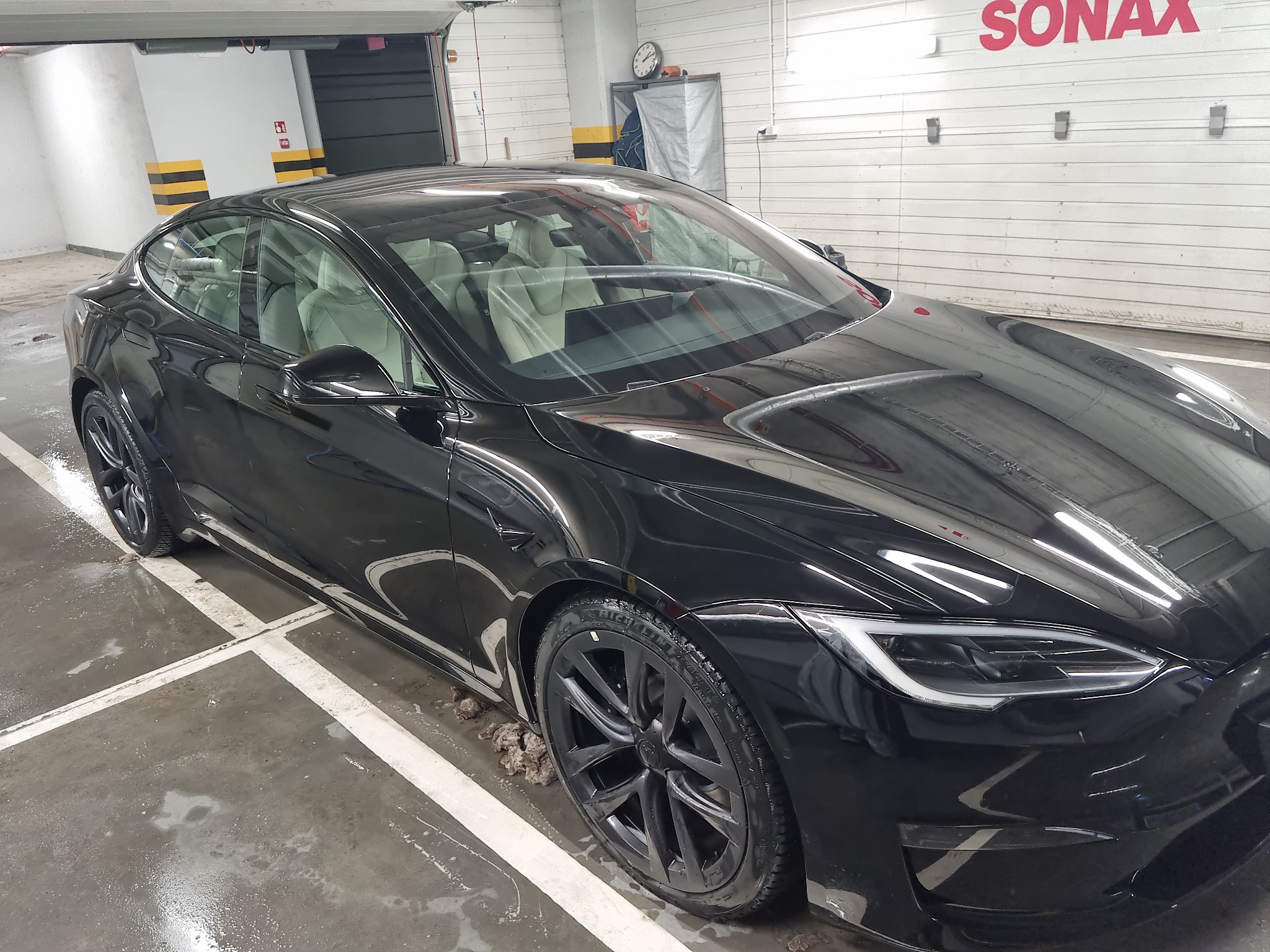 Błyszczący, czarny samochód Tesla Model S po umyciu w myjni; widoczne ślady wody na podłodze i oponach, logo SONAX na ścianie w tle.