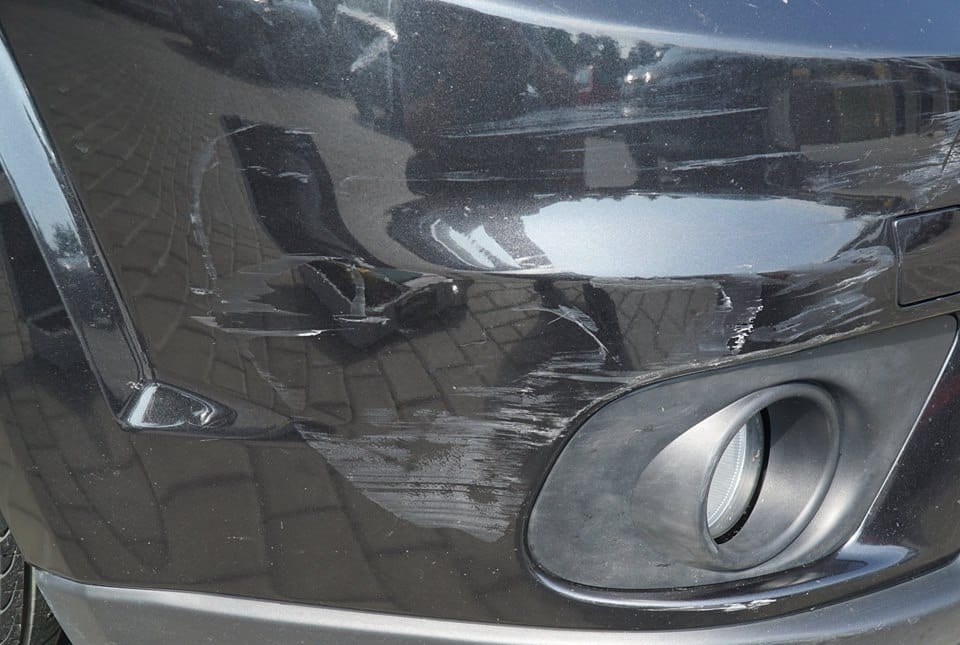 Uszkodzony, porysowany, czarny przedni zderzak samochodu z widocznym światłem przeciwmgielnym.