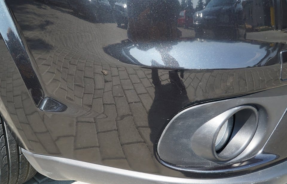 Błyszczący, czarny zderzak samochodu po umyciu, odbijający kostkę brukową i sylwetkę fotografa.