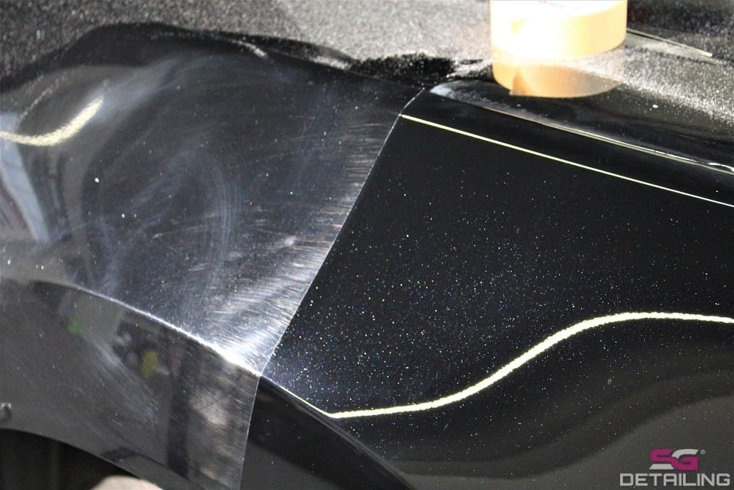 Część karoserii czarnego, metalicznego samochodu, z widoczną różnicą w połysku i stanie lakieru przed i po polerowaniu, z taśmą maskującą oddzielającą obszary.