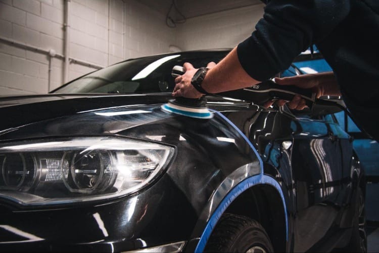 Polerowanie lakieru czarnego samochodu przy użyciu polerki elektrycznej, z niebieską taśmą zabezpieczającą elementy.