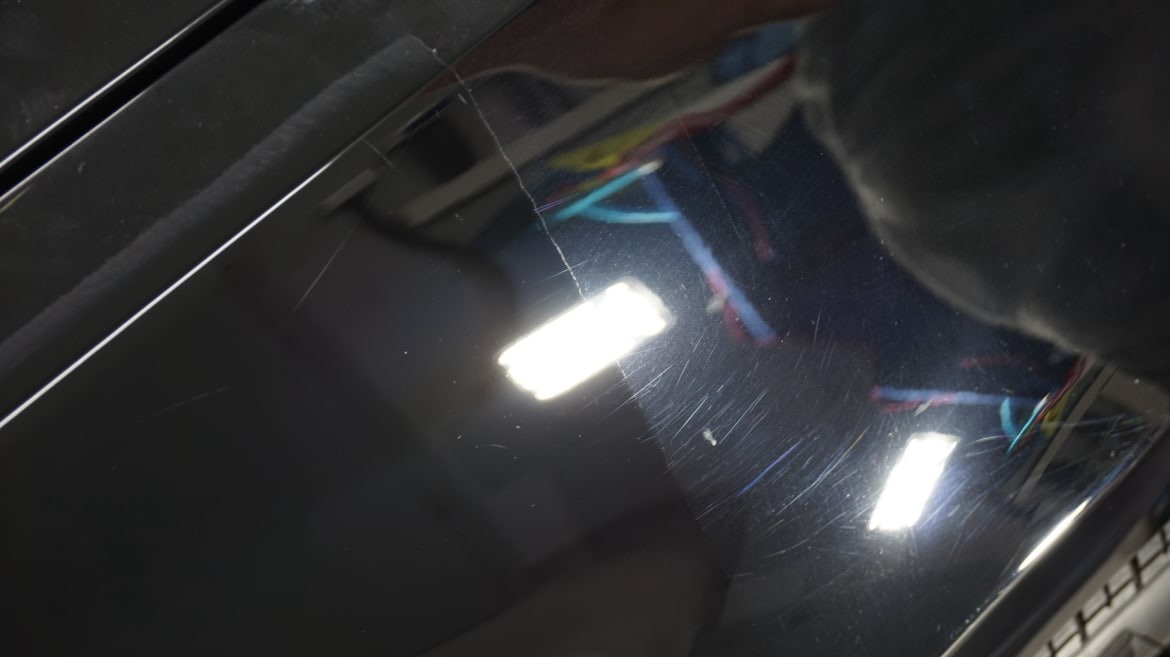 Błyszcząca, ciemna powierzchnia z widocznymi drobnymi rysami i smugami pod światło. W tle widoczne odbicie kabli i fragment osoby.