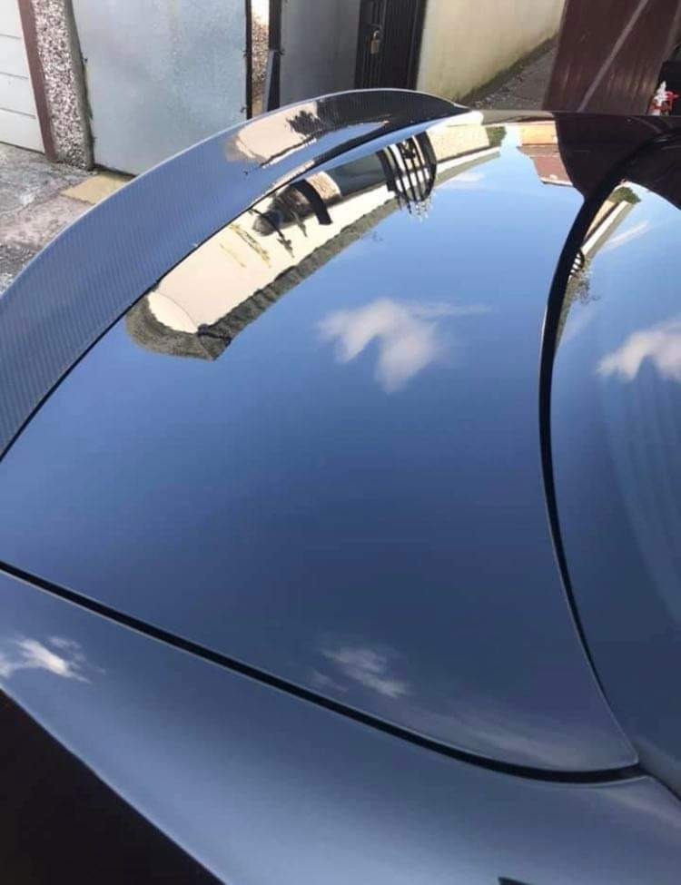 Błyszcząca karoseria czarnego samochodu z karbonowym spojlerem, odbijająca budynek i błękitne niebo z chmurami.