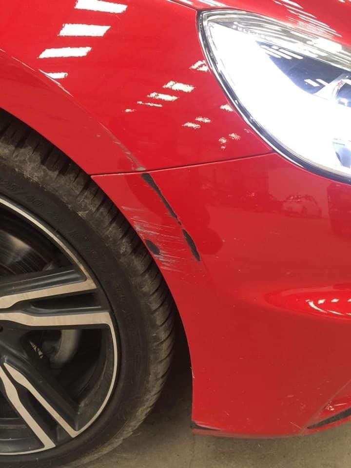 Czerwony samochód z widocznymi rysami i zabrudzeniami na przednim błotniku i zderzaku, obok koła z brudną oponą.