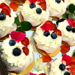 Mini bezy udekorowane bitą śmietaną, borówkami, truskawkami i płatkami róż, ułożone na złotej podkładce na lustrzanej tacy.