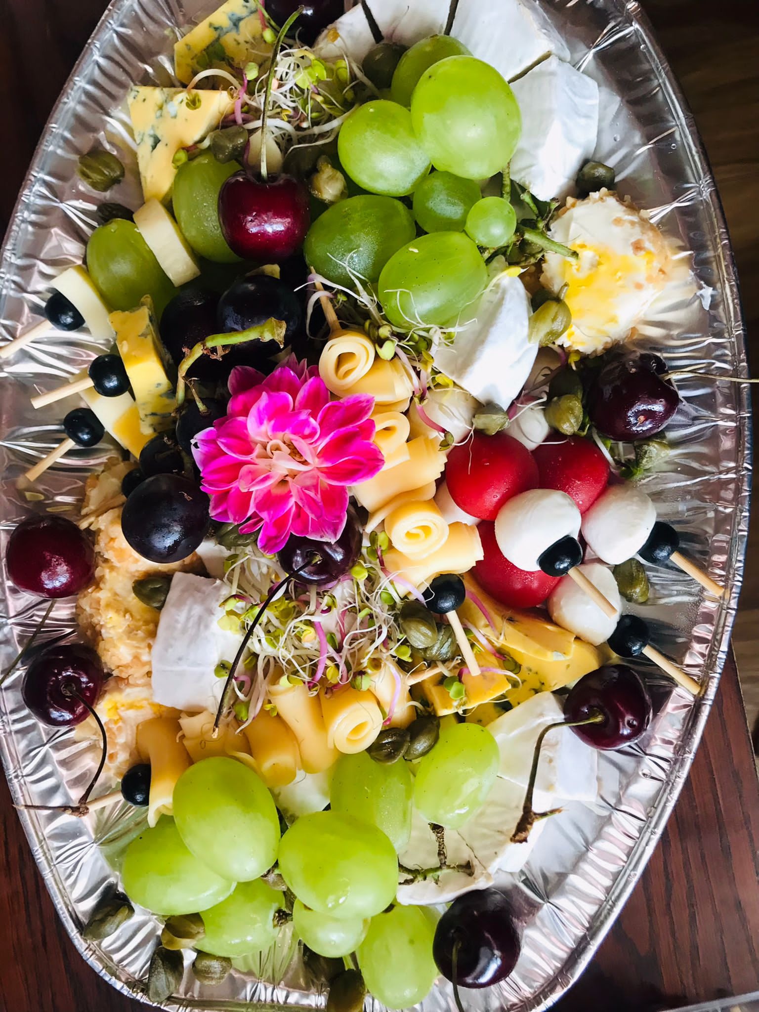 Dekoracyjna srebrna taca z różnorodnym wyborem serów, winogron (zielonych i ciemnych), czereśni, kaparów, kiełków i kwiatu dalii, tworząca apetyczną kompozycję.