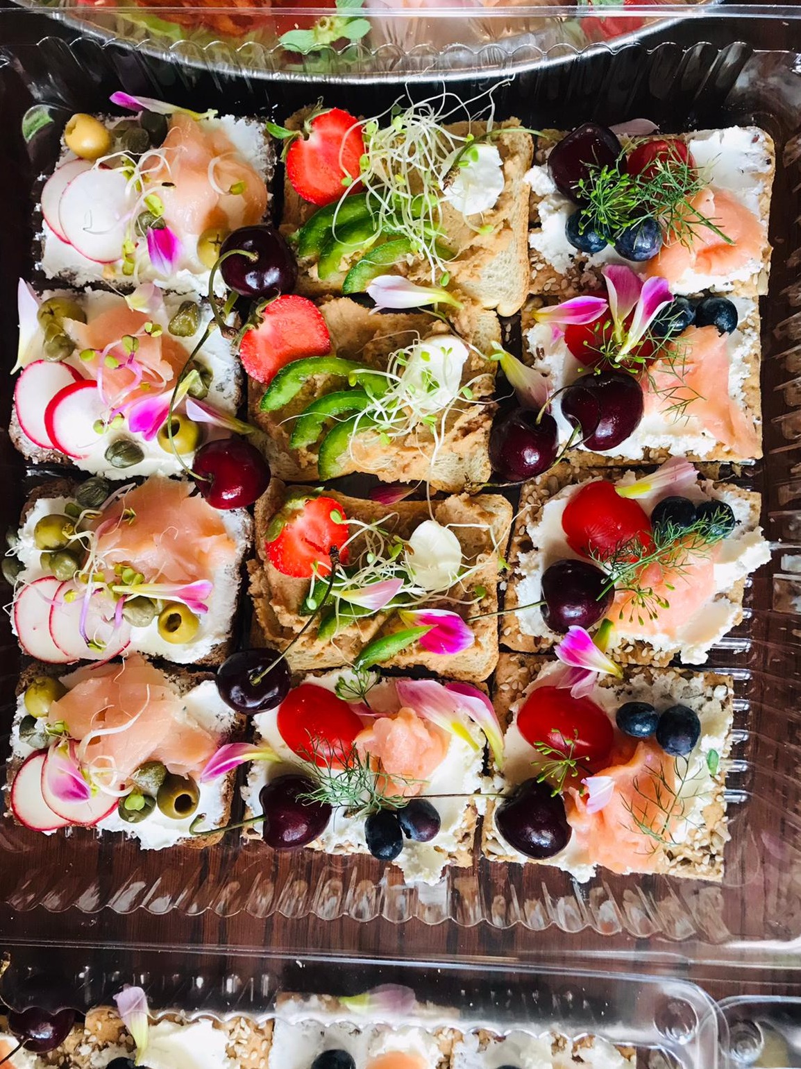 Różnorodne kanapki na krakersach w plastikowym opakowaniu, udekorowane łososiem, serkiem, rzodkiewką, kaparami, oliwkami, truskawkami, zielonymi warzywami, kiełkami, czereśniami, borówkami...