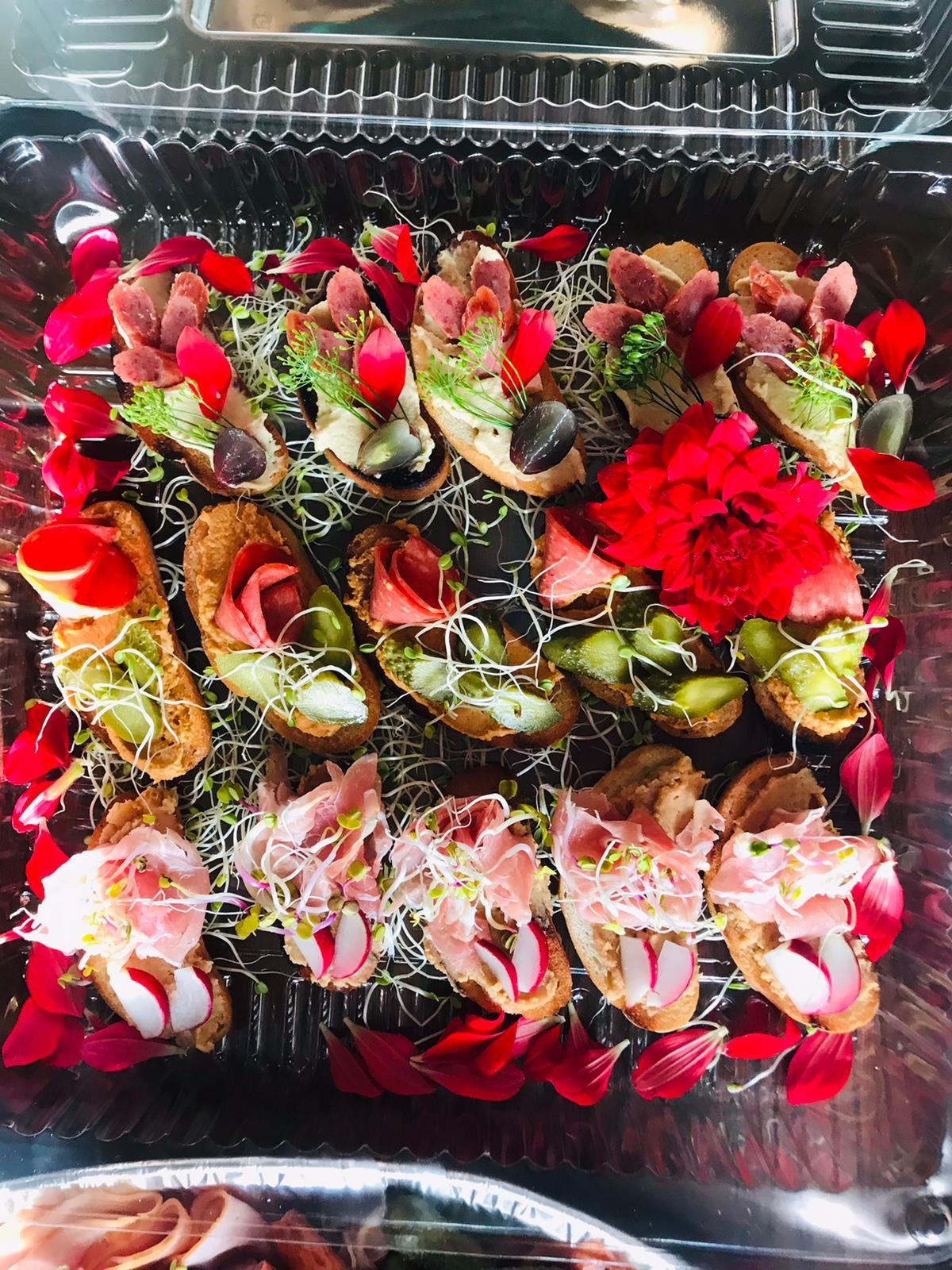 Dekoracyjnie ułożone kanapeczki na pieczywie, z różnymi dodatkami: wędliną, warzywami i kiełkami, ozdobione płatkami czerwonych kwiatów w przezroczystym opakowaniu.