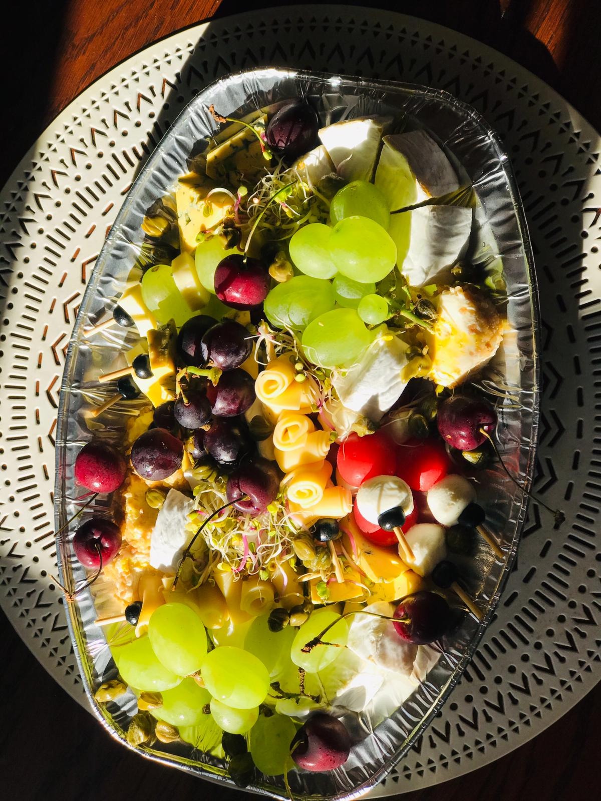 Dekoracyjna taca z serami, winogronami (zielonymi i ciemnymi), czereśniami i mini-szaszłykami z mozzarelli, ułożona na ażurowej podkładce w mocnym, naturalnym świetle słonecznym.