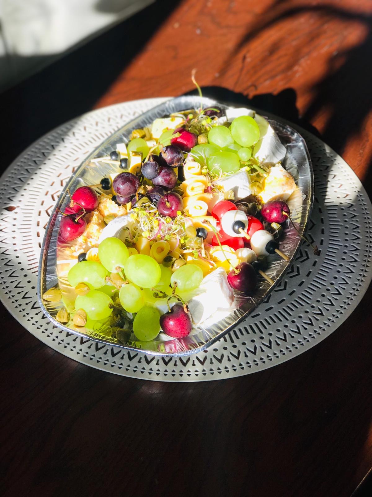 Srebrna taca z kompozycją serów brie, winogron (zielonych i ciemnych), czereśni, pasty w kształcie spirali, pistacji i kiełków, umieszczona na białej, wzorzystej podkładce na ciemnym drewnianym...