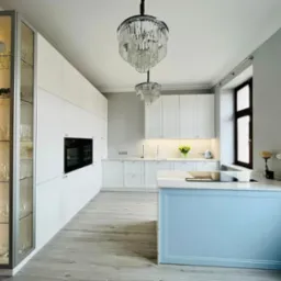 Nowoczesna kuchnia z białymi szafkami, przeszkloną witryną z podświetleniem i kolekcją szkła, wyspą z błękitnym frontem i blatem imitującym marmur oraz dwoma kryształowymi żyrandolami.