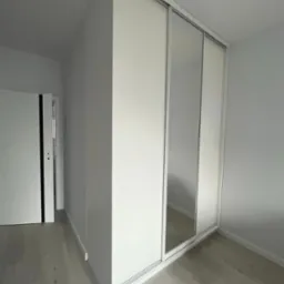 Biała szafa wnękowa z przesuwnymi drzwiami lustrzanymi w jasnym pomieszczeniu, widoczny fragment podłogi i drzwi wejściowe.