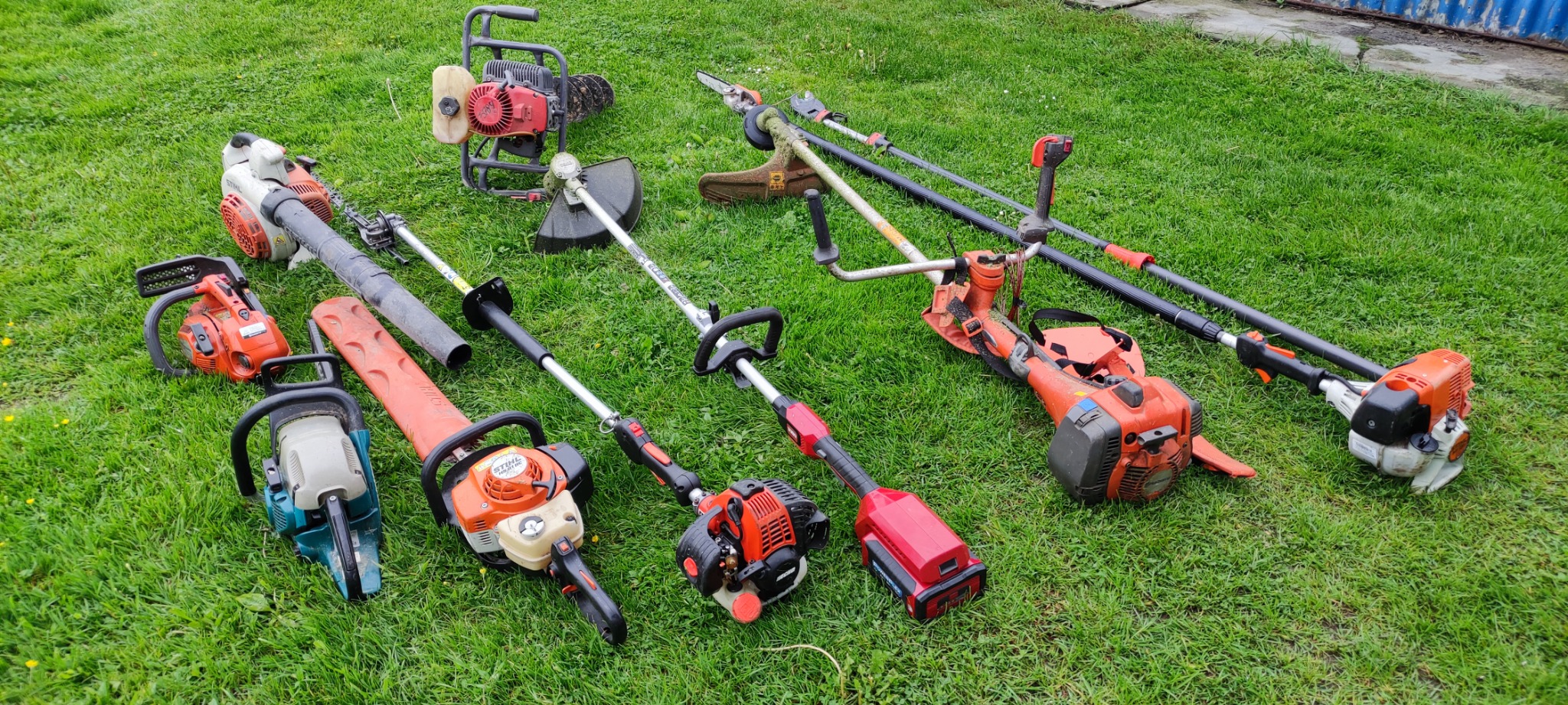 Zestaw różnorodnych urządzeń ogrodniczych, w tym pilarki spalinowe, kosy spalinowe i nożyce do żywopłotu, ułożonych na zielonej trawie. Widoczna różnorodność marek i modeli.