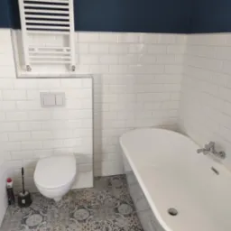 Nowoczesna łazienka z białą wanną wolnostojącą, toaletą podwieszaną, grzejnikiem drabinkowym i wzorzystą podłogą z płytek.