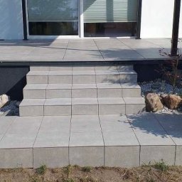 UsługiRemontoweKamil - Schody i taras wyłożone szarą kostką brukową, z widocznym fragmentem ogrodu z roślinami i dekoracyjnymi kamieniami.