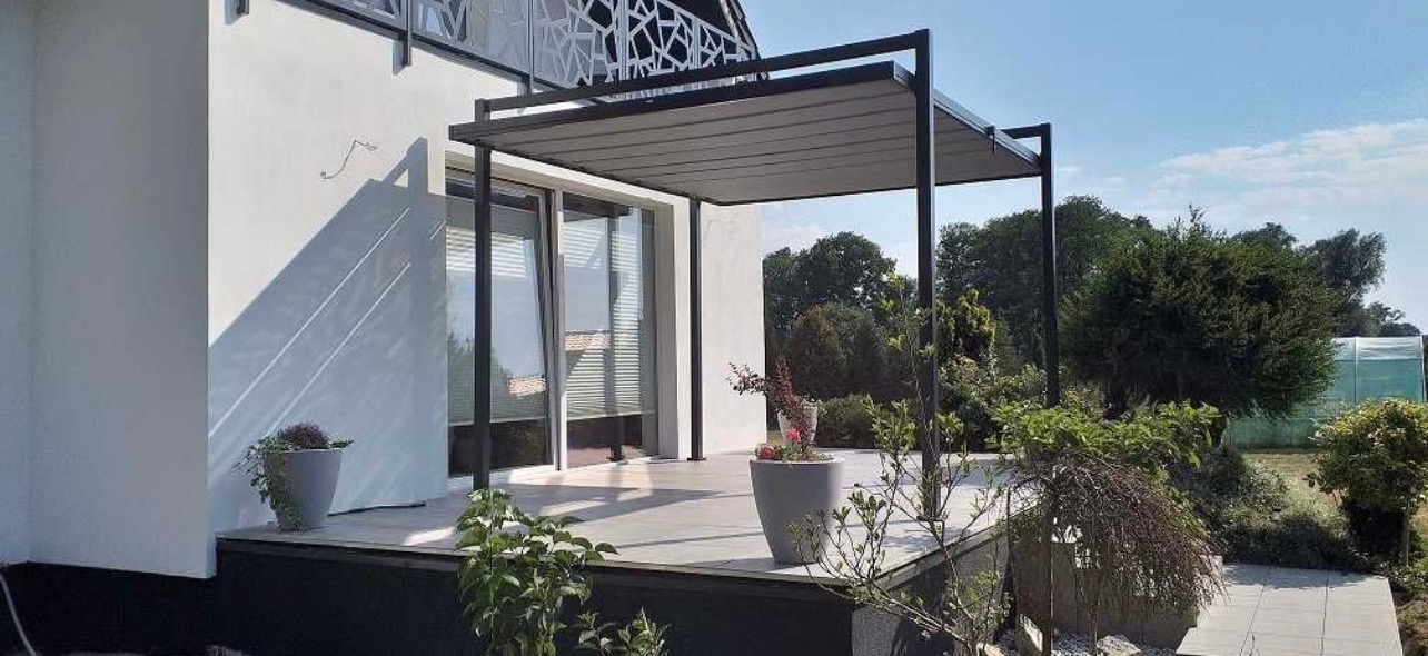 Nowoczesna pergola metalowa z szarym zadaszeniem lamelowym w Pułtusku, przylegająca do budynku z dużymi oknami przesuwnymi, otoczona zielenią i donicami z kwiatami.