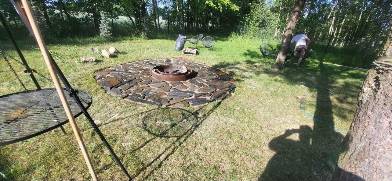 Okrągłe palenisko z kamienia łupanego na trawniku, z metalową misą w środku, obok metalowy grill na trójnogu i krzesła ogrodowe w tle.