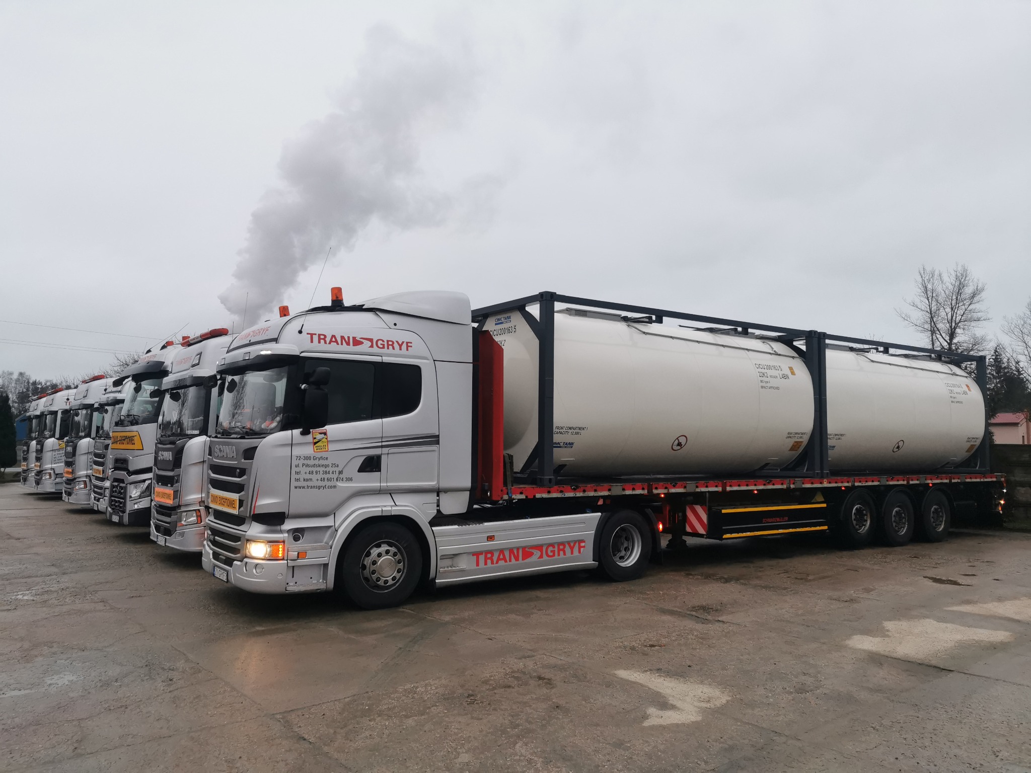 Srebrne ciężarówki marki Scania z logo firmy TRANS-GRYF ustawione w rzędzie, przewożące białe cysterny w kontenerach, w tle komin z dymem.